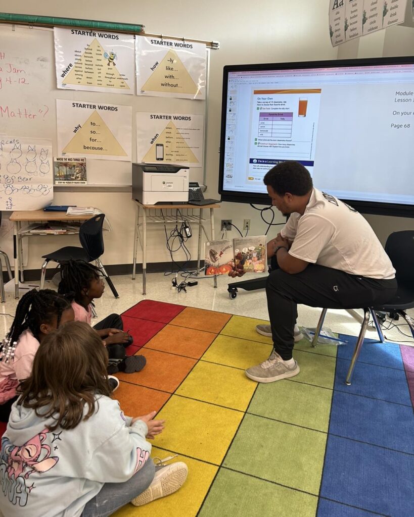 A teacher reads a book to a small group of young students sitting on a colorful classroom rug.