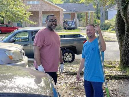 Imagine a scene where two helpful people are standing together, smiling for a photo. One of them is holding a long-handled tool called a rake. In the background, there are five cars parked, and there's a tree on the right side of the photo.