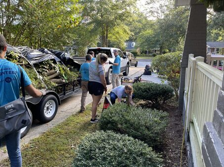 In the picture, a group of volunteers wearing blue shirts is helping to trim bushes at a house with grey painted brick and a white railing. There is also a trailer in the image that is full of branches and limbs.