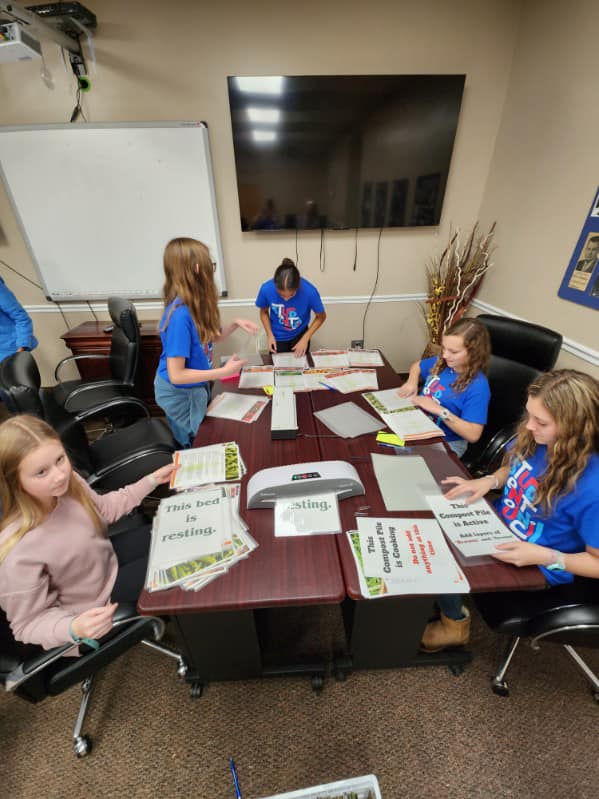 A group of young girls wearing all blue shirts are actively participating in creating a sign as part of the Ready Readers program. In the background, there is a television with wires hanging down, and to the left, there is a smart board. There are all black chairs and four desks arranged together.