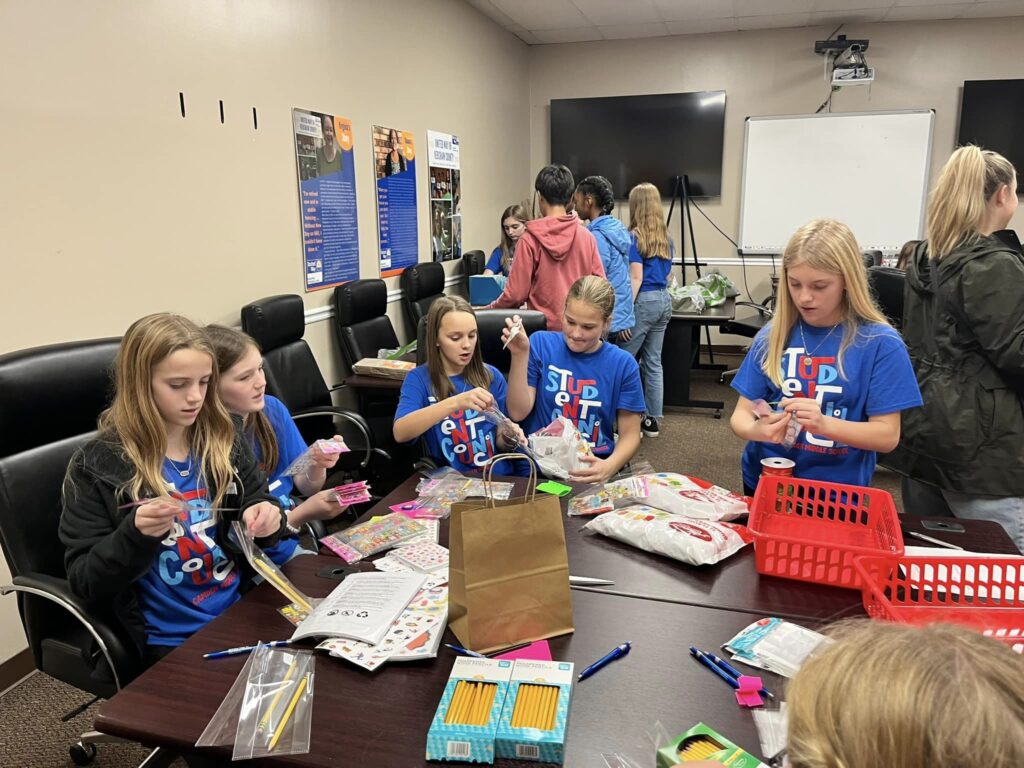 A group of young girls wearing United Way shirts are assisting in organizing and sorting pencils, pens, and sticks. In the background, there are two TVs, a smart board, and a projector in the middle. Along the wall, there is a row of black office chairs. Additionally, there is another group of girls positioned in the back of the room.