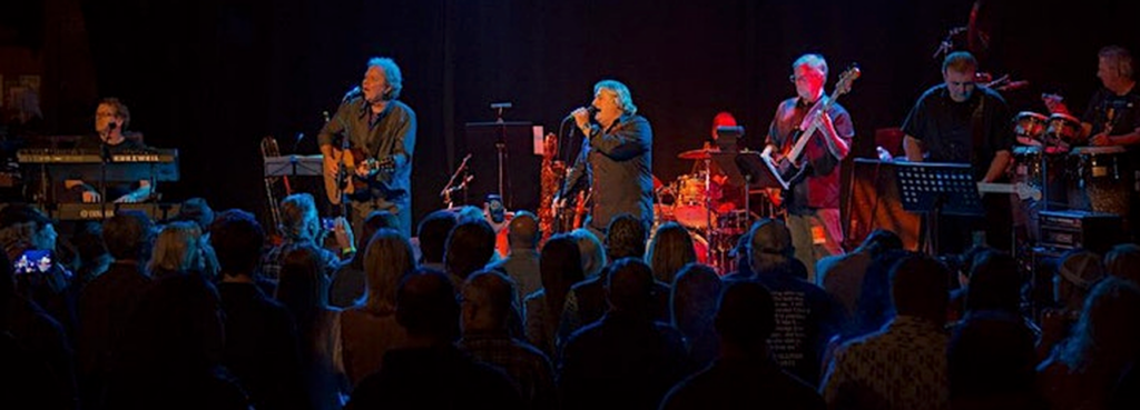 Imagine a special event called the Caring Heart Benefit with a live band performing. There is one person playing the keyboard, guitar, mic, bass, and drums. In the front, there is a crowd of people enjoying the music and the event.