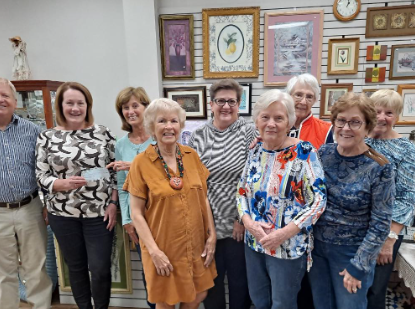 A group of elderly people are standing together, posing for a photo while wearing colorful outfits. In the background, there are many pictures displaying various items.