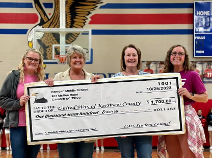 Lori Arledge and three other individuals are pictured holding a check intended for United Way. The photo was taken in front of a school basketball gym that features a logo with an eagle in red and blue.