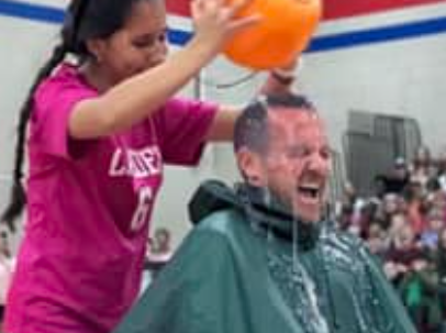 A young girl in a bright pink shirt with black hair is pouring a bucket of water on a man's head. The man is wearing a green poncho, and the bucket is orange. They are in a school gym for a fundraiser.