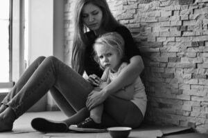 In the picture, there is a mother comforting her baby girl. They look sad, and there is an empty bowl and some cardboard on the floor. It seems like they don't have any food.