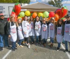 Shows a group of people standing in front of their colorful tent with bright balloons and chili peppers displayed on the side. They are participating in a chili cook-off competition and seem excited about it.