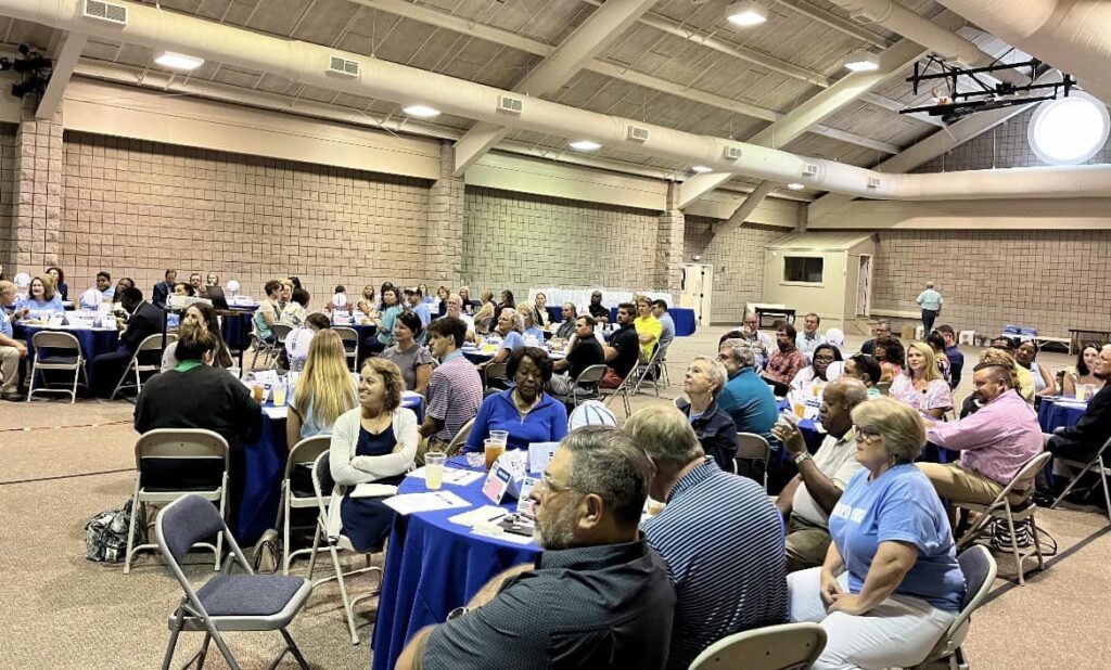 Imagine you are in a large room where a group of people has gathered. There is a speaker talking to everyone, and the room is filled with the sound of their voice. There are several tables in the room covered with blue cloths, and the chairs are made of metal. This creates a setting where many people are seated and focused on listening to the speaker at these tables.