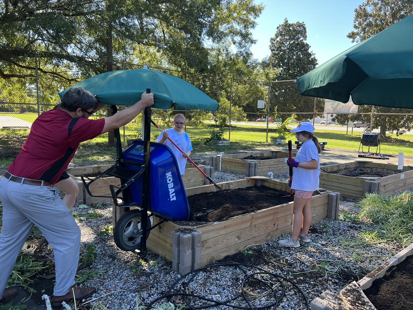 A group of people are working together to build garden boxes for the community. There is an adult using a wheelbarrow to pour soil into the garden box, and two girls are spreading the soil around to make sure it's evenly distributed.