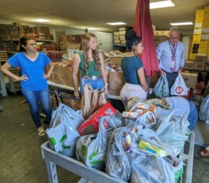 A group of kind volunteers are working together to carry food and provide assistance to the homeless. They are currently located in a warehouse filled with an abundance of food.