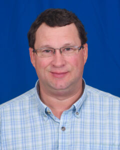 Maury West is a fair-skinned person with rosy cheeks. He is wearing a blue patterned collared shirt and glasses.