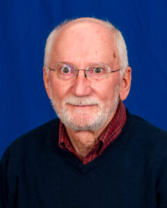 Woody Howland is dressed in a black crewneck sweater with a red collared shirt underneath. He has a white, close-shaved beard and wears glasses. He appears to be an older gentleman.