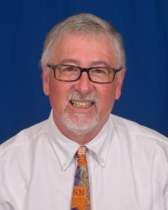 Dan Mathews is wearing a white shirt with a tie that represents Clemson University. He is an older gentleman who wears glasses and has a goatee, which is a small beard on the chin.