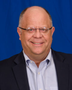 Doug Stone is a mature man who is dressed formally in a light purple dress shirt and a sports jacket. He is also wearing glasses.