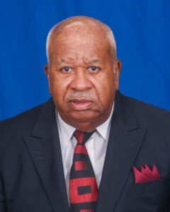 Quinten Chevis is a well-dressed older man with a mustache. He is wearing a black suit with a white shirt and a black and red tie.