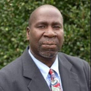 A black man is wearing a grey suit, accented by a blue shirt and a red and blue tie.