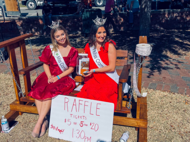 Imagine two women named Miss Camden and Miss Camden Teen, both wearing red dresses and holding a jar filled with money. There's a sign nearby that says "raffles."