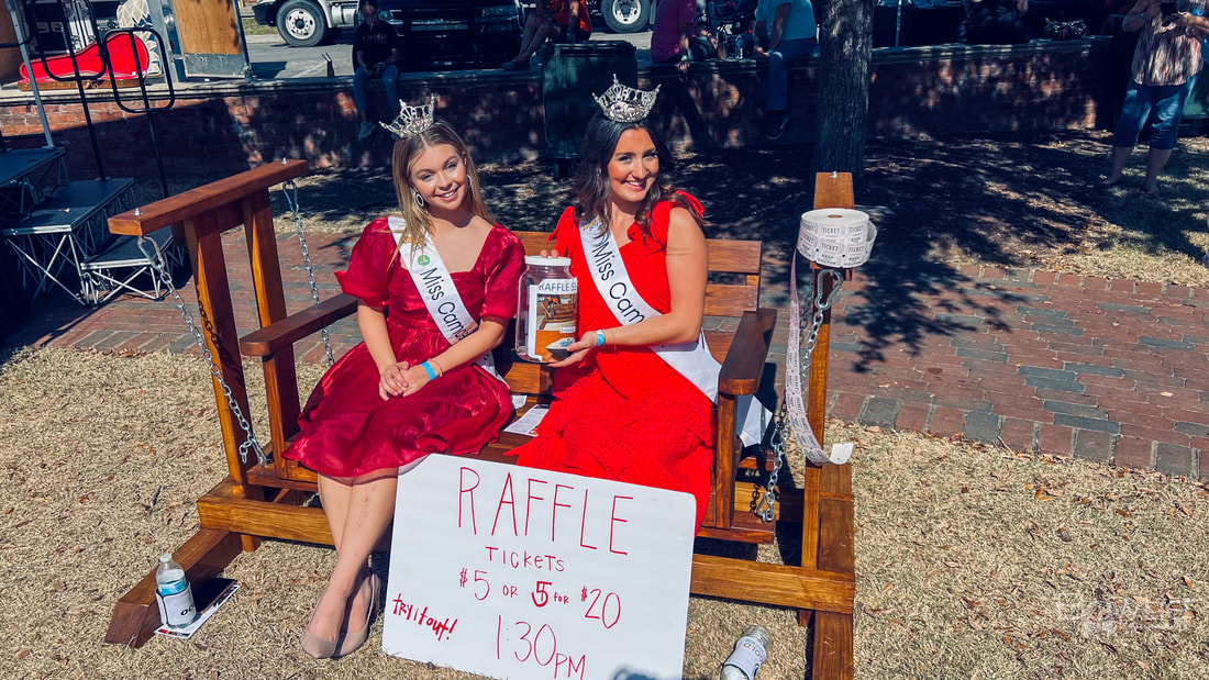 Imagine two women named Miss Camden and Miss Camden Teen, both wearing red dresses and holding a jar filled with money. There's a sign nearby that says "raffles."