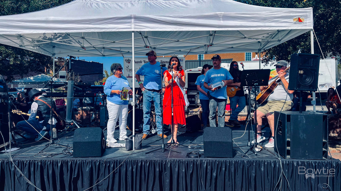 Miss Camden and the United Way are on a stage with a man playing the guitar. The stage is black and there's a white tent.