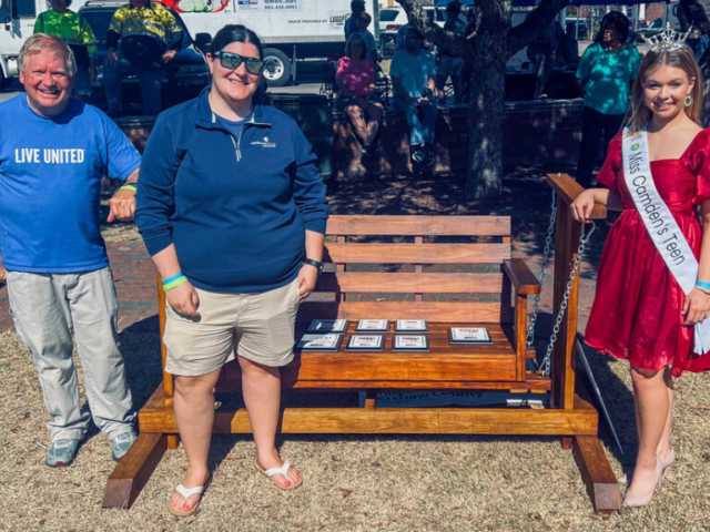 Donny and Miss Camden, both teenagers, are standing in front of a wooden rocking bench with all the awards for the chili contest displayed on it.