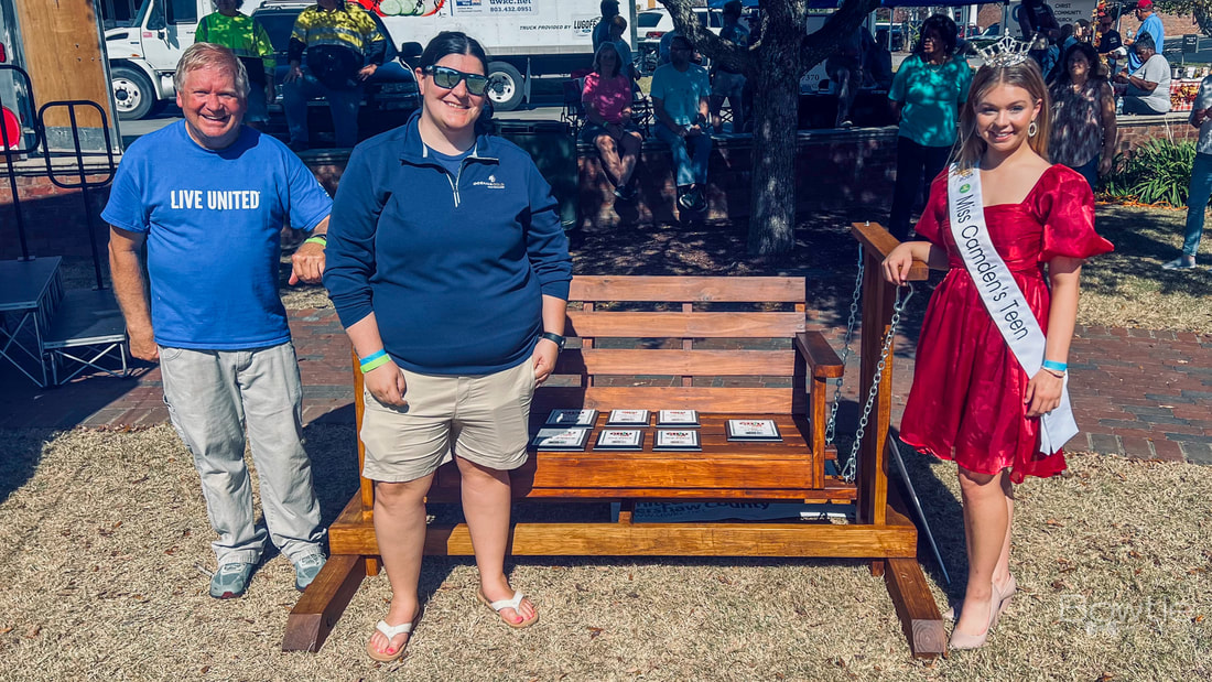 Donny and Miss Camden, both teenagers, are standing in front of a wooden rocking bench with all the awards for the chili contest displayed on it.