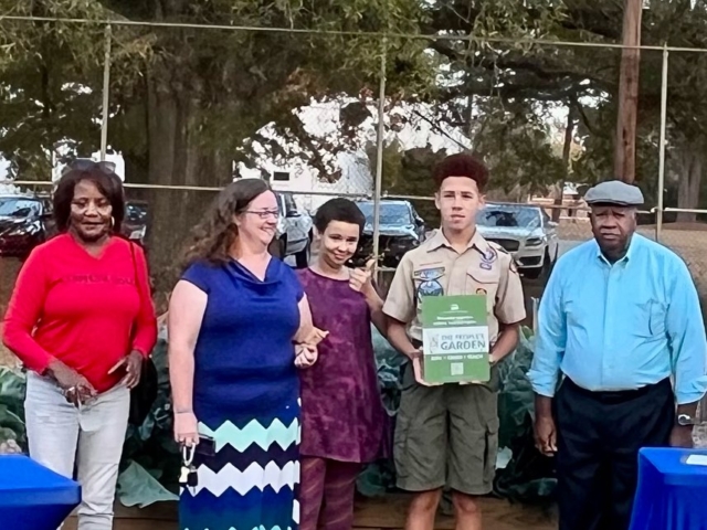 A group of people, including a boy scout and his family, are assembled to take a photo in front of a community garden.