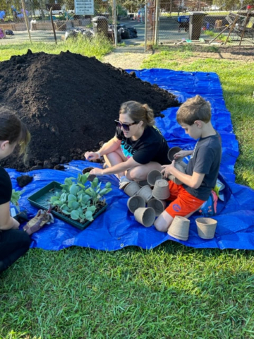 A mother and her children are working together to plant seeds in a plot, so they can later transfer the plants to the community garden. They are currently seated next to a large pile of dirt, with the community garden visible in the background.