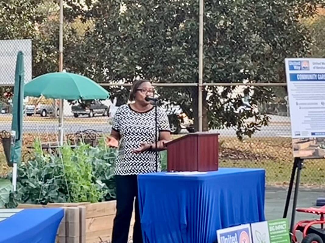 Imagine a woman standing confidently and speaking to a group of people. Behind her, there are raised garden boxes filled with green plants. The woman is sharing her words, while the beauty of nature surrounds her.