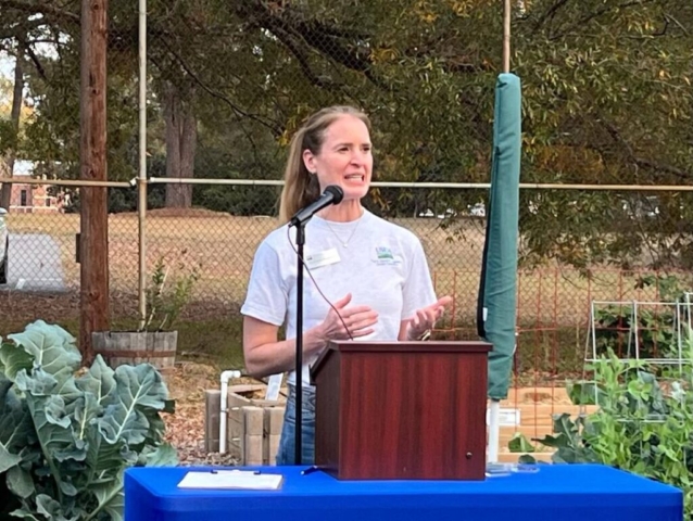 Imagine a woman standing confidently and speaking to a group of people. Behind her, there are raised garden boxes filled with green plants. The woman is sharing her words, while the beauty of nature surrounds her.