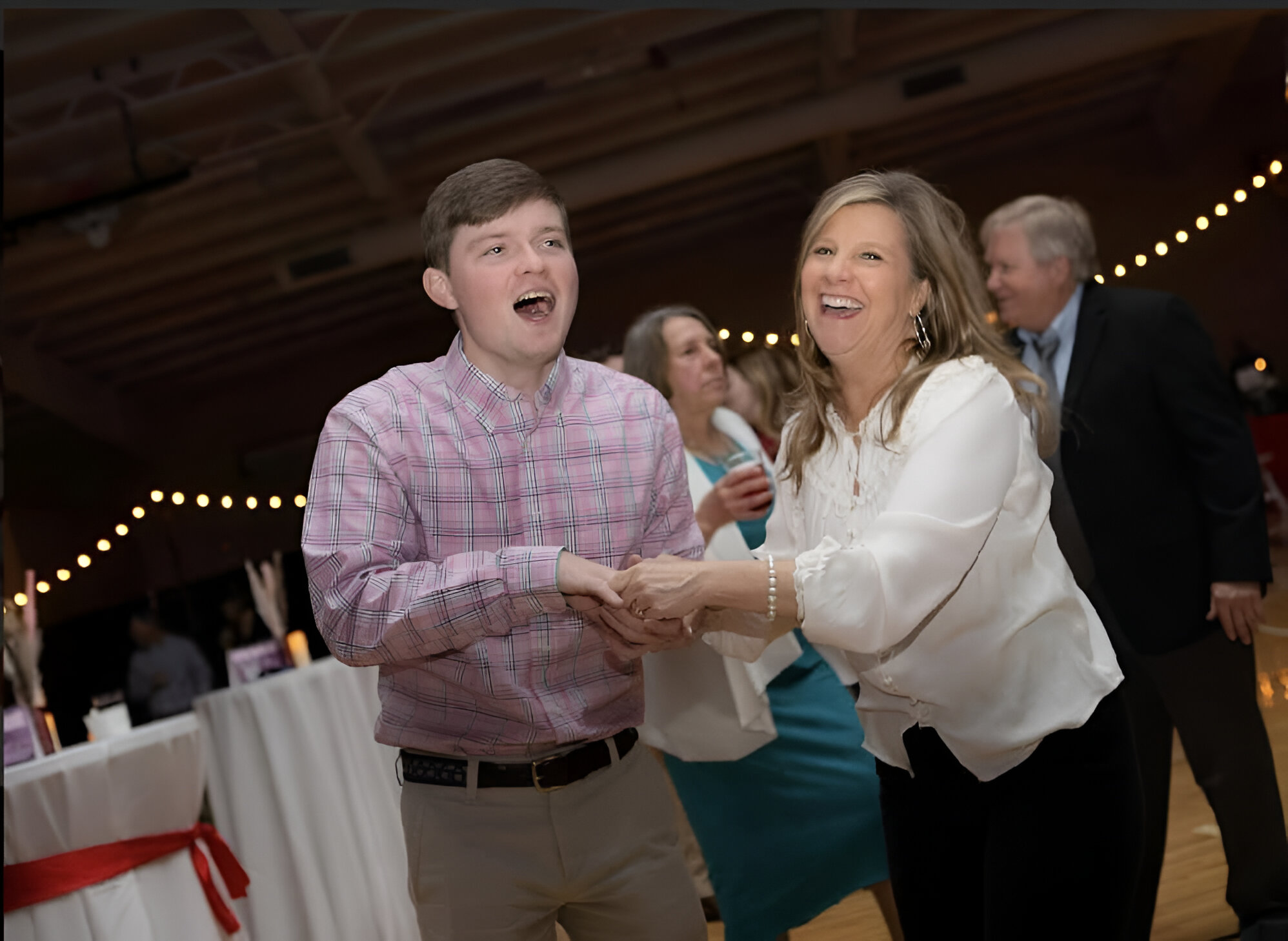 There is a group of people in the background who are dressed up and dancing. In the forefront, there is a mother and her son dancing together. The son is wearing a pink dress shirt and khakis, while the mother is wearing a white shirt with black pants.