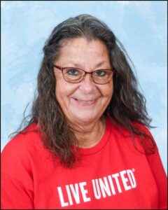 There is a woman who is older and has long grey hair. She is wearing a red shirt with the words "Live United" and she wears glasses.