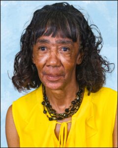 A woman with dark skin and black hair that goes to her shoulders. She is wearing a necklace and has a yellow shirt on.