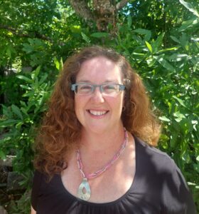 A woman who is older and has long curly hair. She is wearing a black shirt and she wears glasses. Also, she is wearing a necklace around her neck.