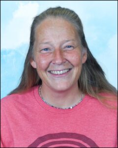 A person with long grey hair. She is wearing a small necklace and a pink shirt.