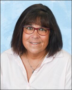 A woman wearing a white dress shirt. She is also wearing a necklace and red glasses. Additionally, she has shoulder-length dark hair.