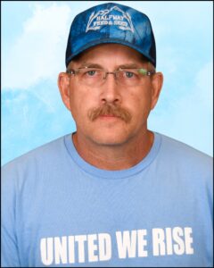 A man wearing a light blue shirt with the words "United we rise" written on it. He is older, has a blonde mustache, wears glasses, and is wearing a blue hat with a logo that seems to be a combination of the words "fred" and "seed."