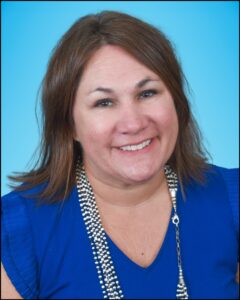 A woman with brown hair that falls to her shoulders. She is dressed in a blue shirt and is wearing a long necklace around her neck.