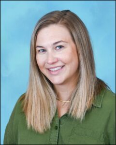 Woman with light blonde and wearing a shiny piece of jewelry around her neck. She has light blue eyes and is dressed in a comfortable green shirt made of soft fabric.