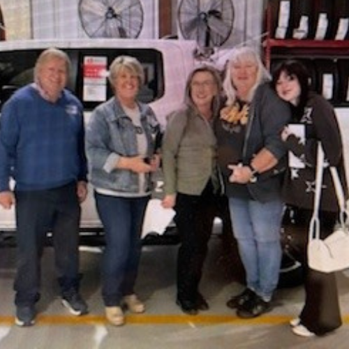 Five adults stand smiling together in front of a white vehicle in an indoor setting.