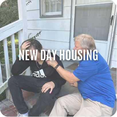 A man in blue comforts another man sitting on porch steps; text reads NEW DAY HOUSING.
