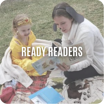 An adult and a child in costume read a book together on a blanket outdoors. Text: Ready Readers.