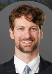 Smiling man with a beard and wavy hair wearing a suit, tie, and white shirt against a gray background.