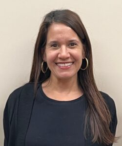A woman with long brown hair, hoop earrings, and a black top smiles in front of a beige wall.