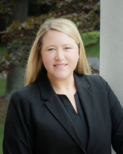 Woman with long blonde hair in a black blazer, standing outdoors by a light-colored wall, smiling.