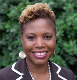 Smiling woman with short curly hair, wearing a brown blazer and earrings, standing outdoors with greenery behind her.