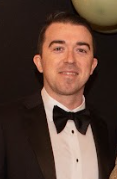 Man in a black tuxedo and bow tie smiling in front of a black background.