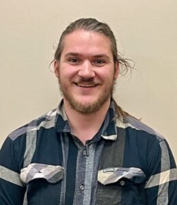 A smiling person with long hair and a beard wearing a plaid shirt stands against a plain beige background.