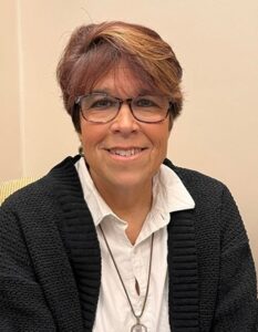 Smiling woman with short brown hair, glasses, and a black sweater over a white shirt, sitting indoors.