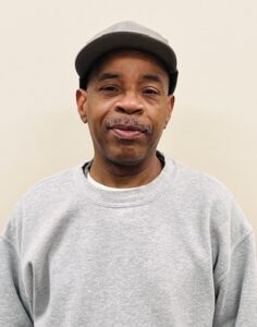 Man wearing a gray sweatshirt and cap standing against a plain light-colored background, looking at the camera.