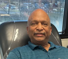 Smiling man in a blue shirt sitting on a chair by a window, with cars and trees visible outside.
